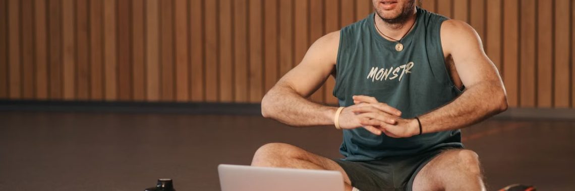 Adult man exercising indoors with a laptop for virtual fitness training, sitting on a red mat.